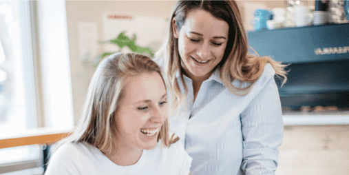 Two women are smiling and appear to be engaged in a friendly conversation while standing indoors at Jeffrey Studios. One woman has blonde hair and wears a white shirt, and the other has lighter brown hair and wears a light blue shirt. They are near a window and a counter, perhaps discussing their favorite Tasmanian website designer.